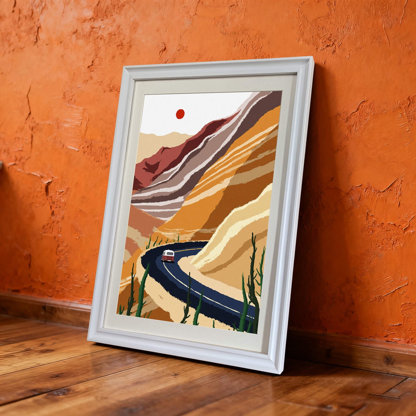 The framed desert road art print in a white wooden frame, leaning on a wooden floor against a vibrant, textured orange wall.