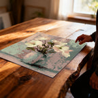 Unframed art print of soft white flowers in a pale vase against a pastel pink and green textured background, lying on a large, rustic wooden table with a person's hand reaching toward it.