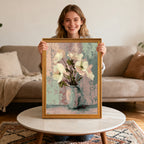 Smiling woman sitting and holding an art print of soft, white flowers in a blue vase with a gold-colored wood frame, centered over a round coffee table.