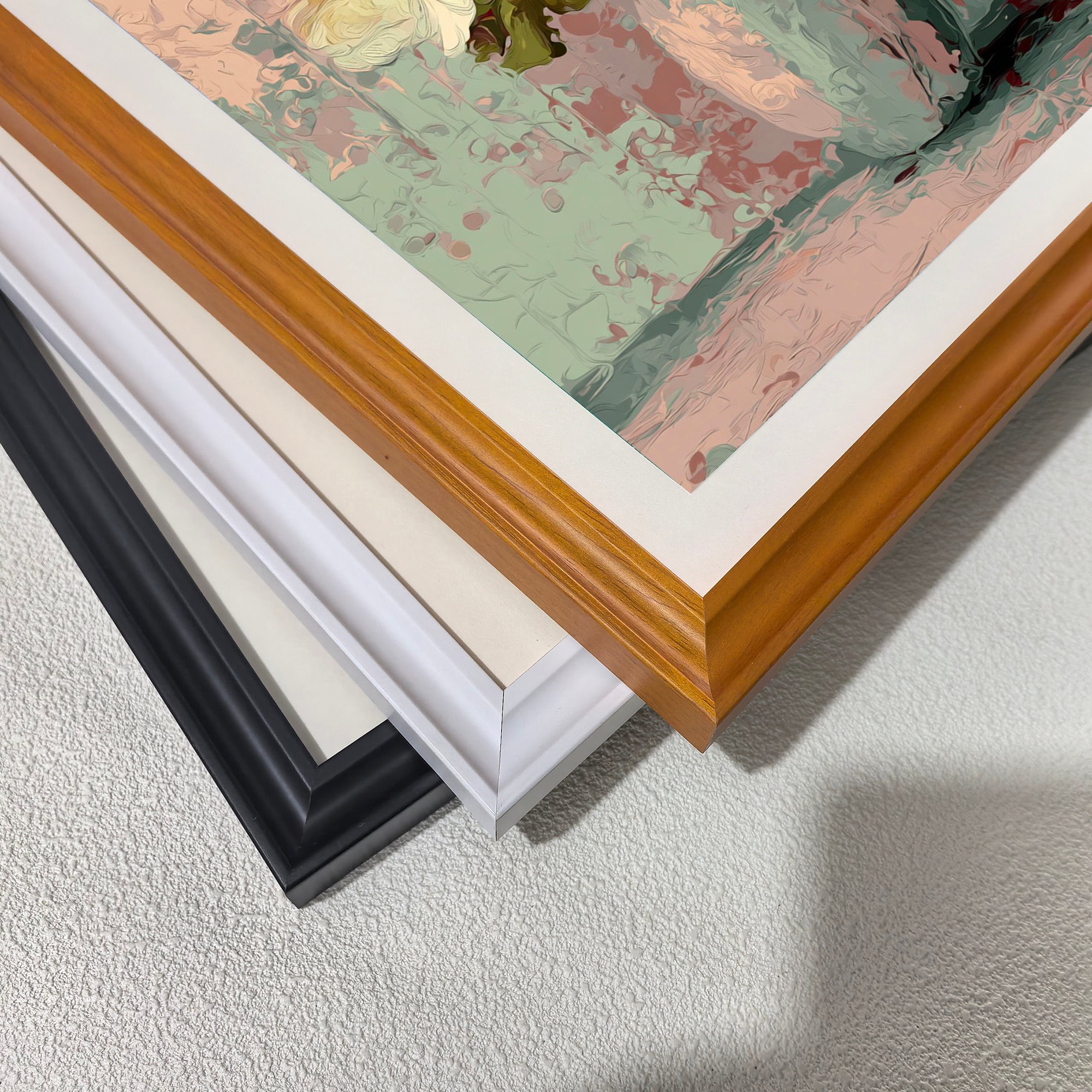 Close-up of three stacked wood picture frame corners in black, white, and natural light brown, showing the art print inside with pastel pink and green colors.
