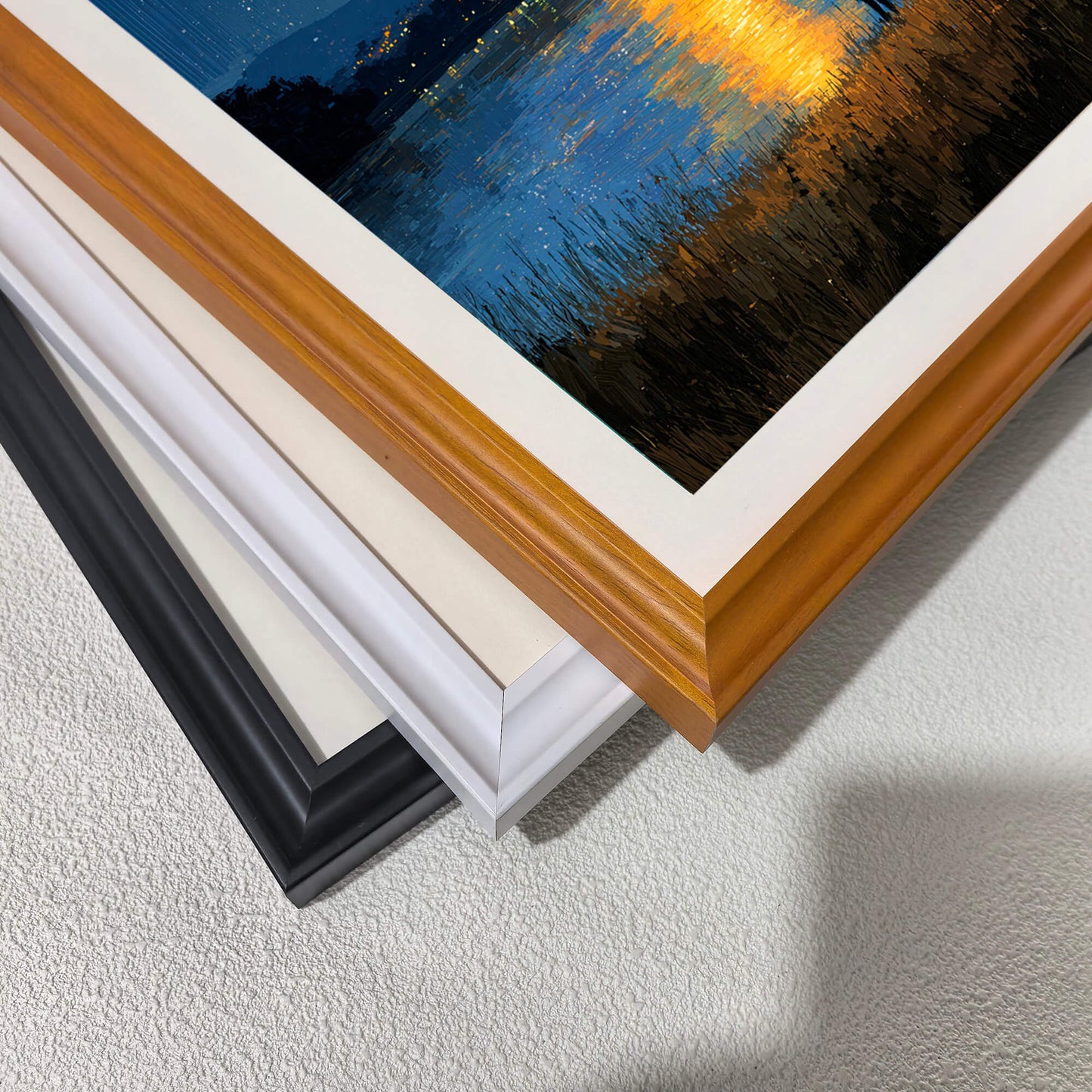 A stack of wood picture frame corners showing different color options: natural wood, cream/off-white, white, and black. A corner of the moonlit lake art print, showing the textured brushwork, is visible on top.