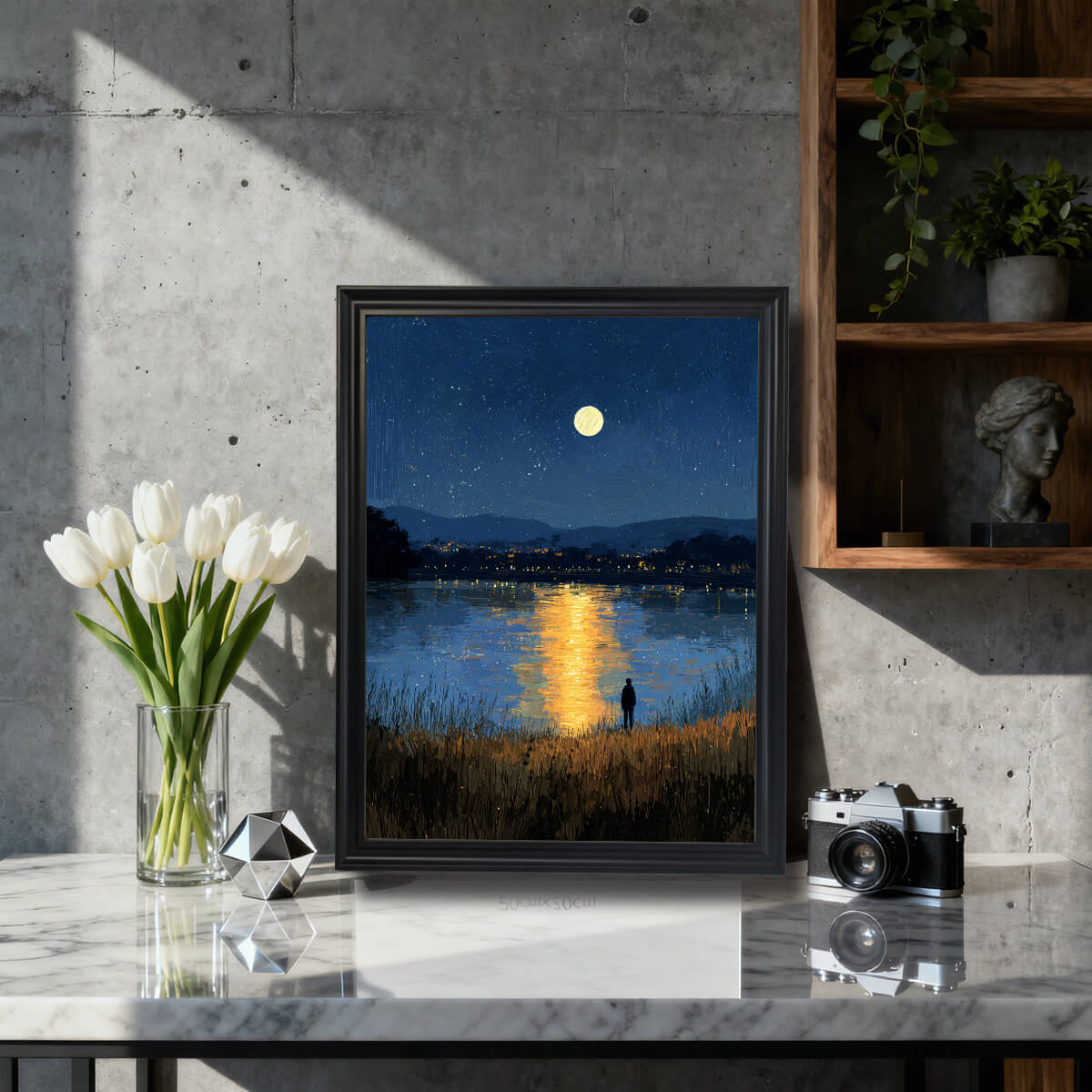 A framed art print of a moonlit, starry night over a lake with distant city lights and a solitary figure in the foreground. The print is in a black frame, resting on a marble-top table next to white tulips and a vintage camera in a modern, concrete-walled setting.