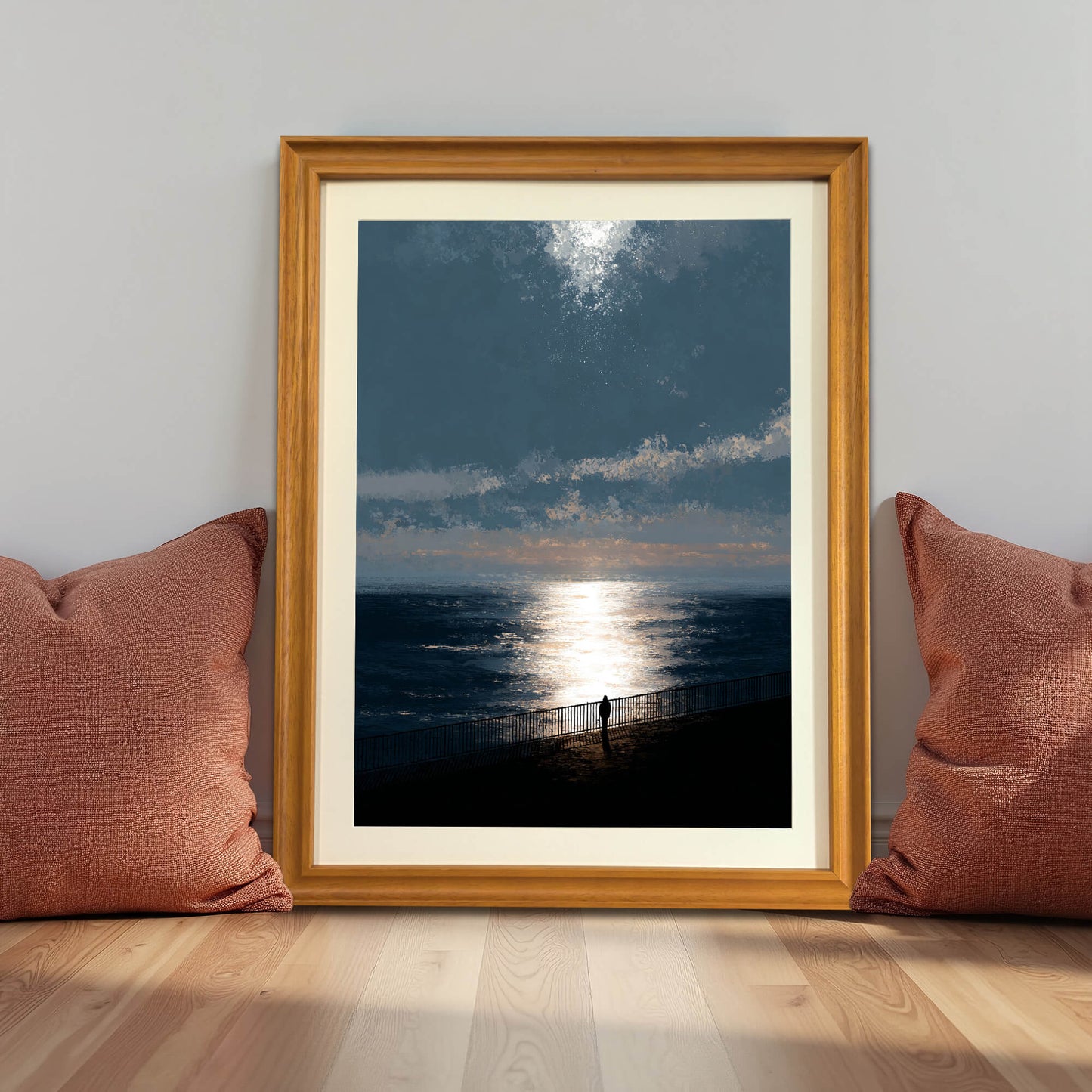 Moody ocean sunset art print in a natural light wood frame (oak/maple), leaning against a gray wall on the floor between two terracotta-colored pillows, demonstrating a cozy, grounded decor style.
