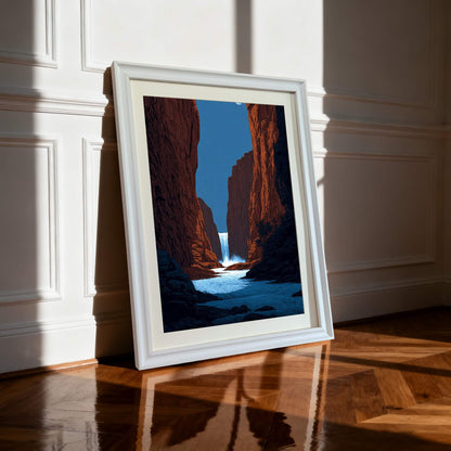 A framed art print of a striking red canyon with a central waterfall and river, leaning against a white wainscoted wall on a glossy hardwood floor, brightly lit by sunlight. The frame is white.