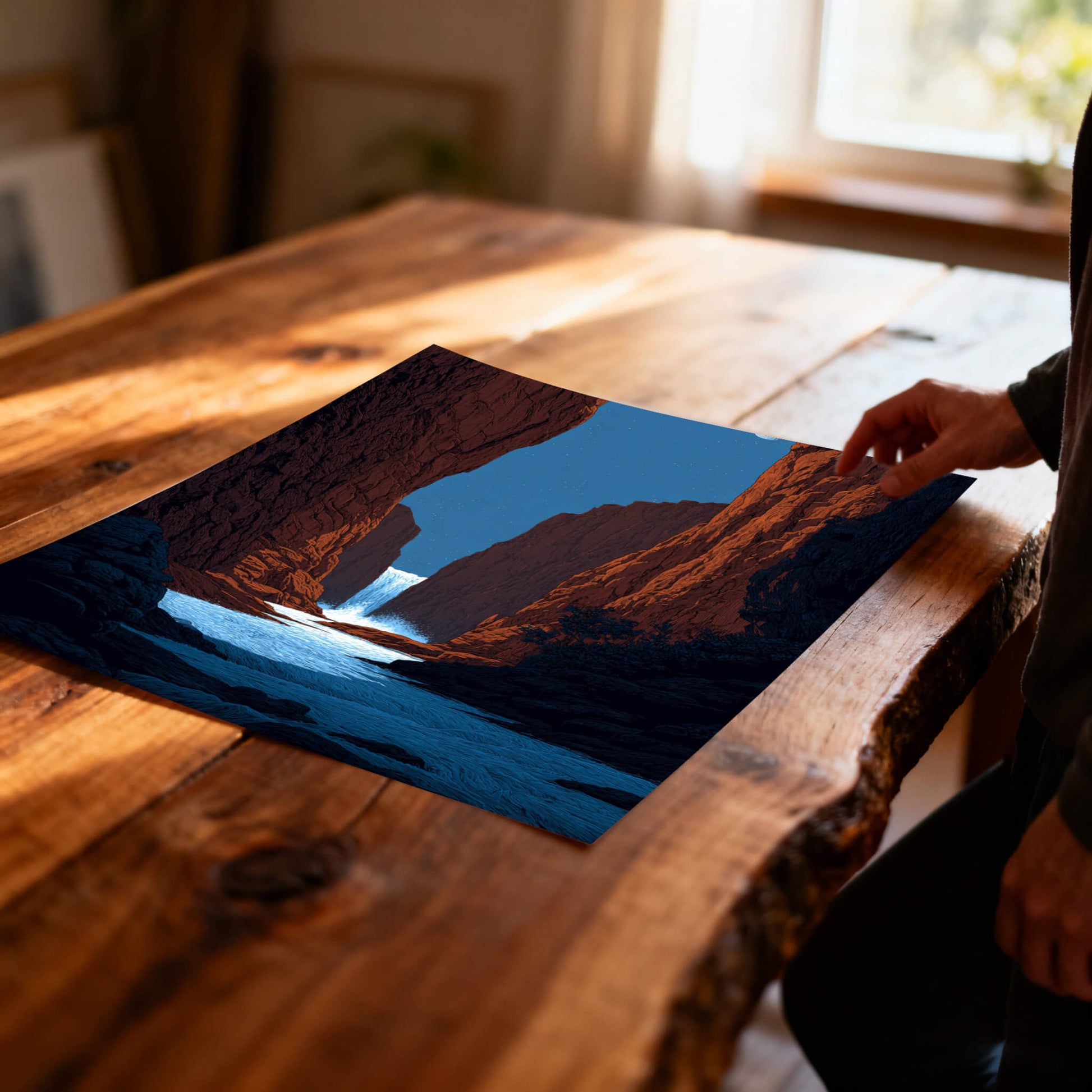 A close-up, angled view of an unframed art print lying on a rustic wooden table. The print shows a dramatic red canyon with a waterfall and a blue river flowing out. A person's hand is visible on the right.