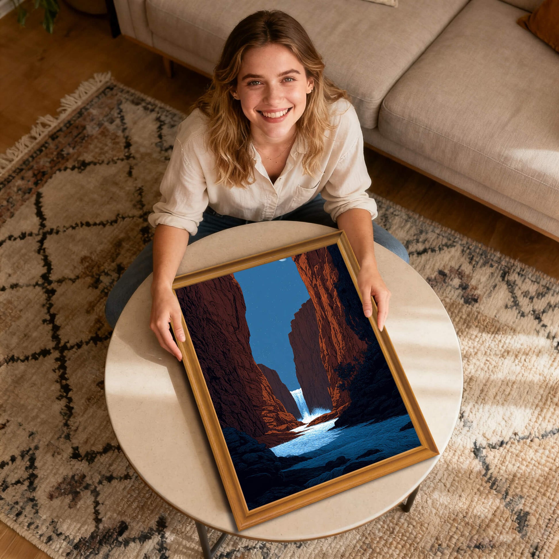 A smiling woman sits on the floor holding a framed art print of a red rock canyon and waterfall under a night sky. The frame is natural wood and rests on a round coffee table on a patterned rug.