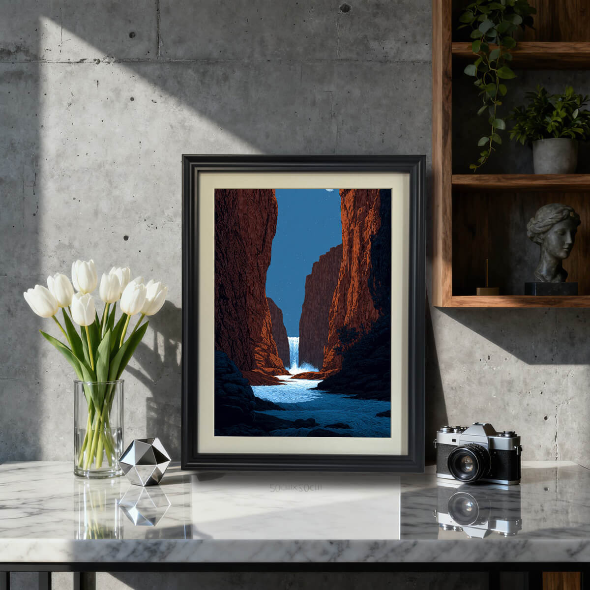 A framed art print depicting a vibrant red rock canyon with a waterfall cascading into a blue river under a starry night sky. The print is in a black frame and is displayed on a marble-top table next to white tulips in a modern, concrete-walled setting.