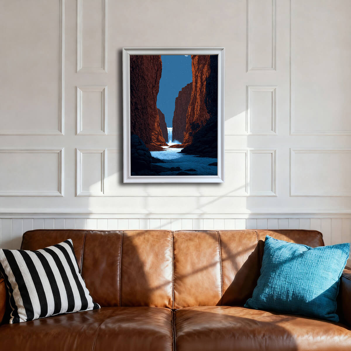 A framed art print of a canyon waterfall under a starry sky, hanging on a white wainscoted wall above a brown leather sofa. The frame is white.