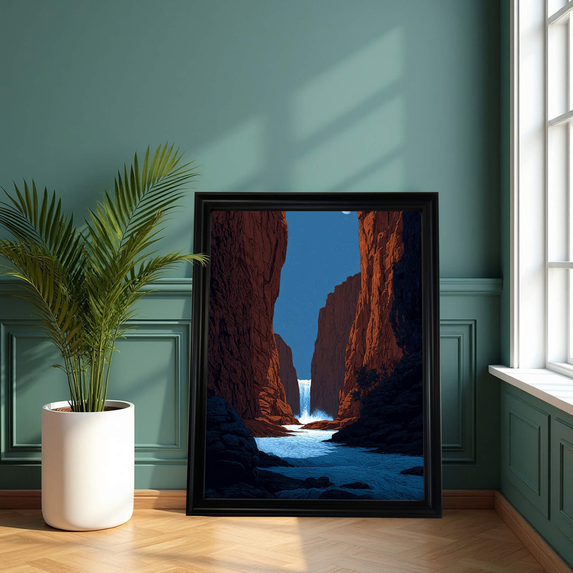 A framed art print of a dramatic red rock canyon and waterfall, resting on a wooden floor against a dark, paneled green wall, beside a tall potted palm plant. The frame is black.