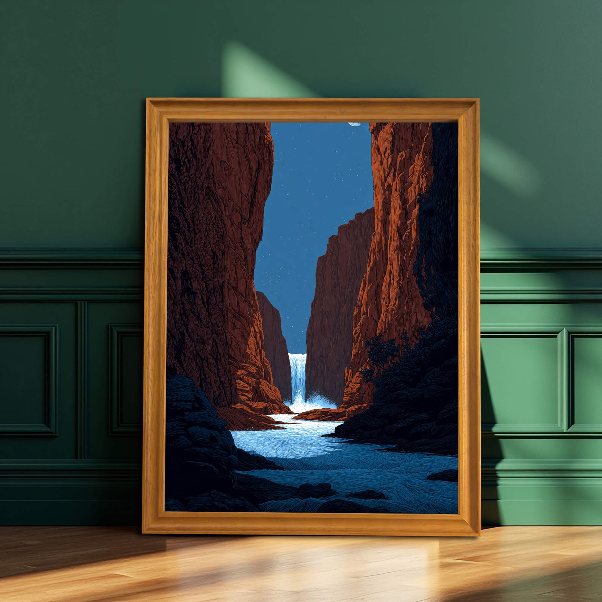 A framed art print of a vibrant red canyon with a waterfall and blue river, resting on a wooden floor against a dark, paneled green wall. The frame is natural wood/light brown.
