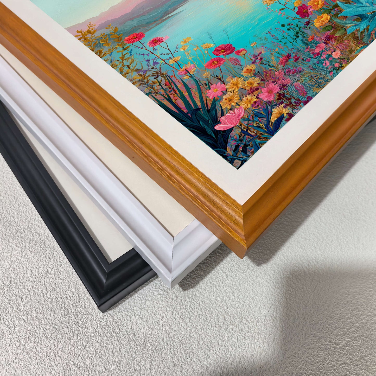 A close-up, top-down view of three picture frames (black, white, and wood-toned) stacked diagonally on a white textured surface, showcasing the different frame finishes.