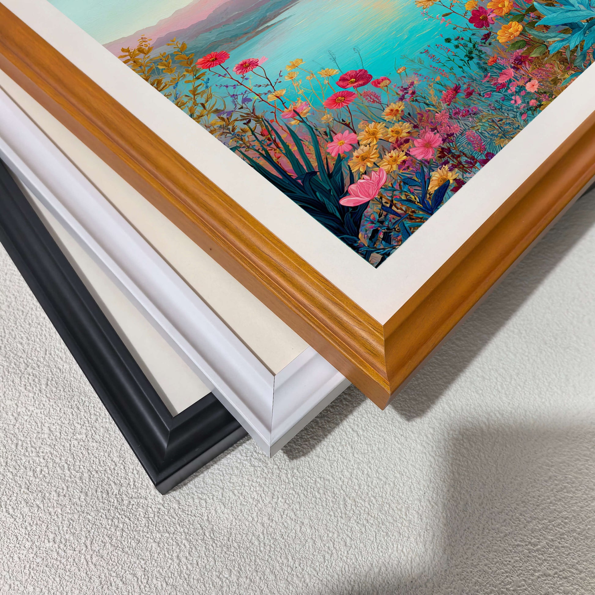 A close-up, top-down view of three picture frames (black, white, and wood-toned) stacked diagonally on a white textured surface, showcasing the different frame finishes.