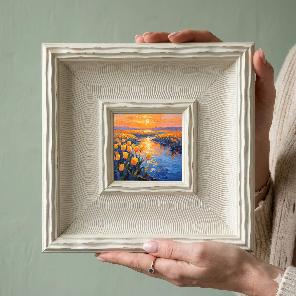 A small framed painting depicting a sunset over a field of yellow tulips and a blue river. The painting is in a white, textured frame. The artwork is held in front of a light green background.