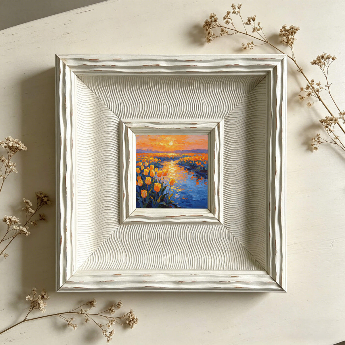 A small, framed painting of a sunset over a field of yellow flowers and a blue river. The painting is set within a white, textured frame. Dried flowers are placed around the frame.