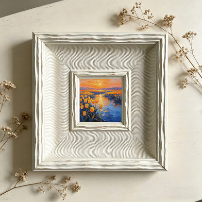  A small, framed painting of a sunset over a field of yellow flowers and a blue river. The painting is set within a white, textured frame. Dried flowers are placed around the frame.
