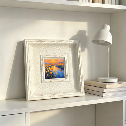 A square, white framed painting with a landscape scene of yellow flowers and a river at sunset. The frame has a textured, wavy design. A white desk lamp and stacked books are also visible.