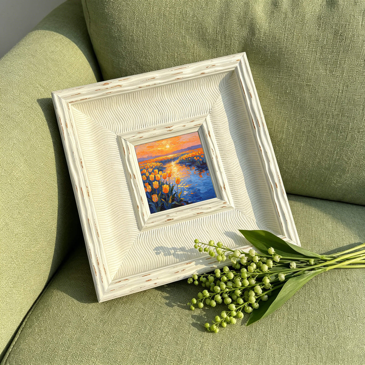 A framed oil painting of a sunset over a field of yellow flowers and a river. The painting is in a white, textured frame. A bouquet of lily of the valley is in the foreground. The painting is on a green surface.