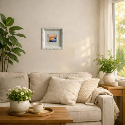A small, square painting of a sunset scene, framed in a distressed white wooden frame. The painting depicts a vibrant sky with orange and blue hues. The artwork is displayed on a neutral-colored wall above a sofa.