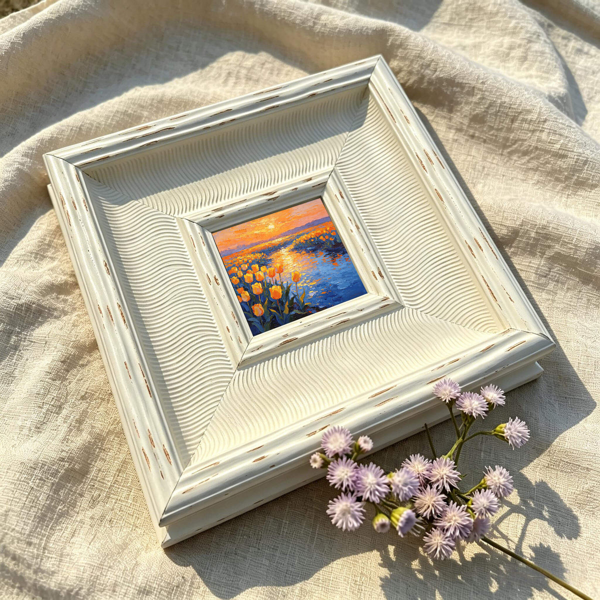 A small framed painting with a landscape scene of yellow flowers and a body of water under an orange sunset. The frame is off-white with a textured, wavy design. A sprig of purple flowers rests near the frame.