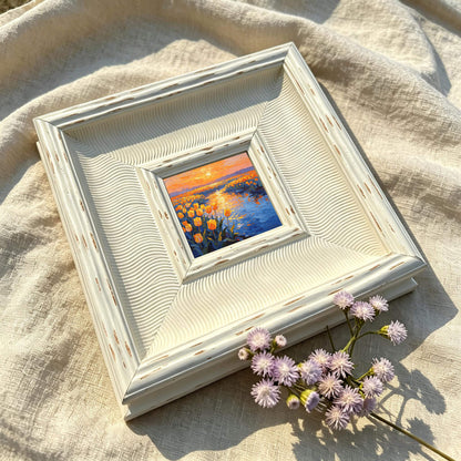 A small framed painting with a landscape scene of yellow flowers and a body of water under an orange sunset. The frame is off-white with a textured, wavy design. A sprig of purple flowers rests near the frame.
