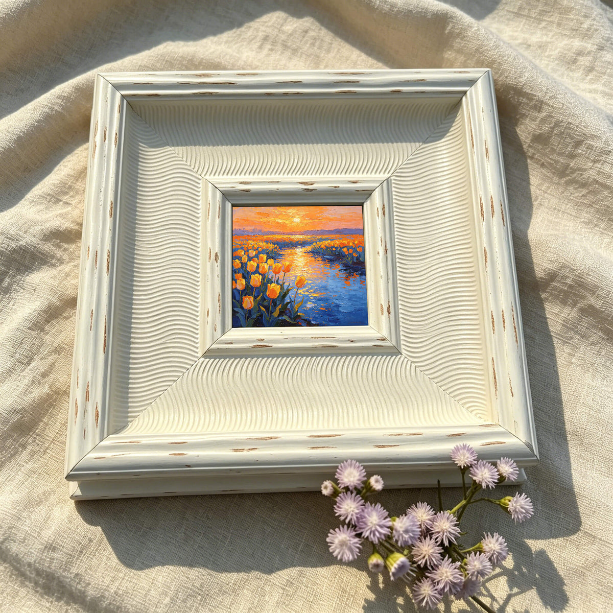 Square, off-white framed painting of a sunset over a body of water and yellow flowers. The painting is centered within the frame, which has a textured, wavy design. A small bouquet of purple flowers rests in the lower right corner.