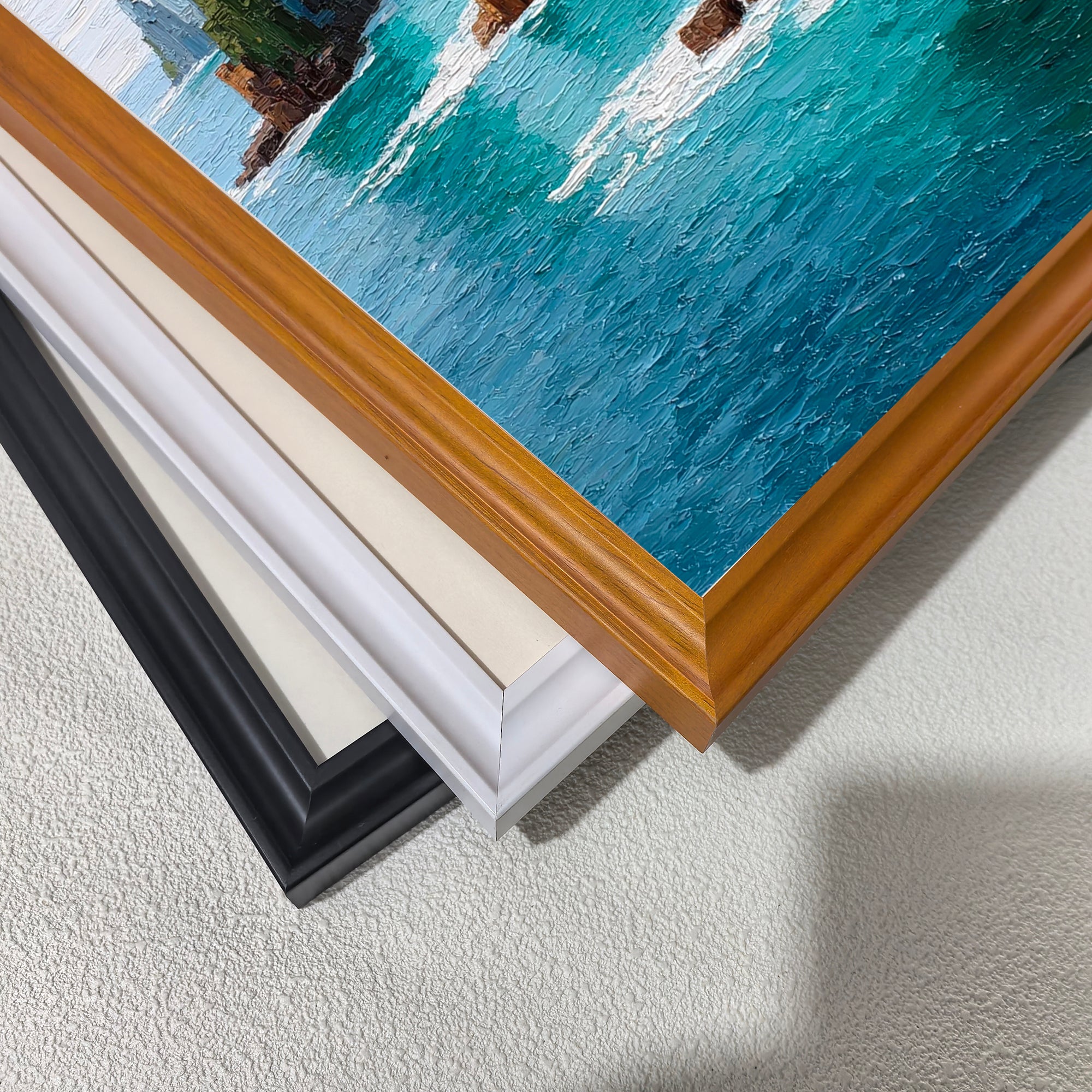 A corner close-up showing the detail of the textured oil painting of ocean waves and cliffs, with four different frame and mat options stacked: warm wood, white, cream, and black.