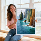 A smiling woman sits on a window sill, holding the art print framed in a sleek, silver or light grey frame, showcasing the artwork next to a bright window.