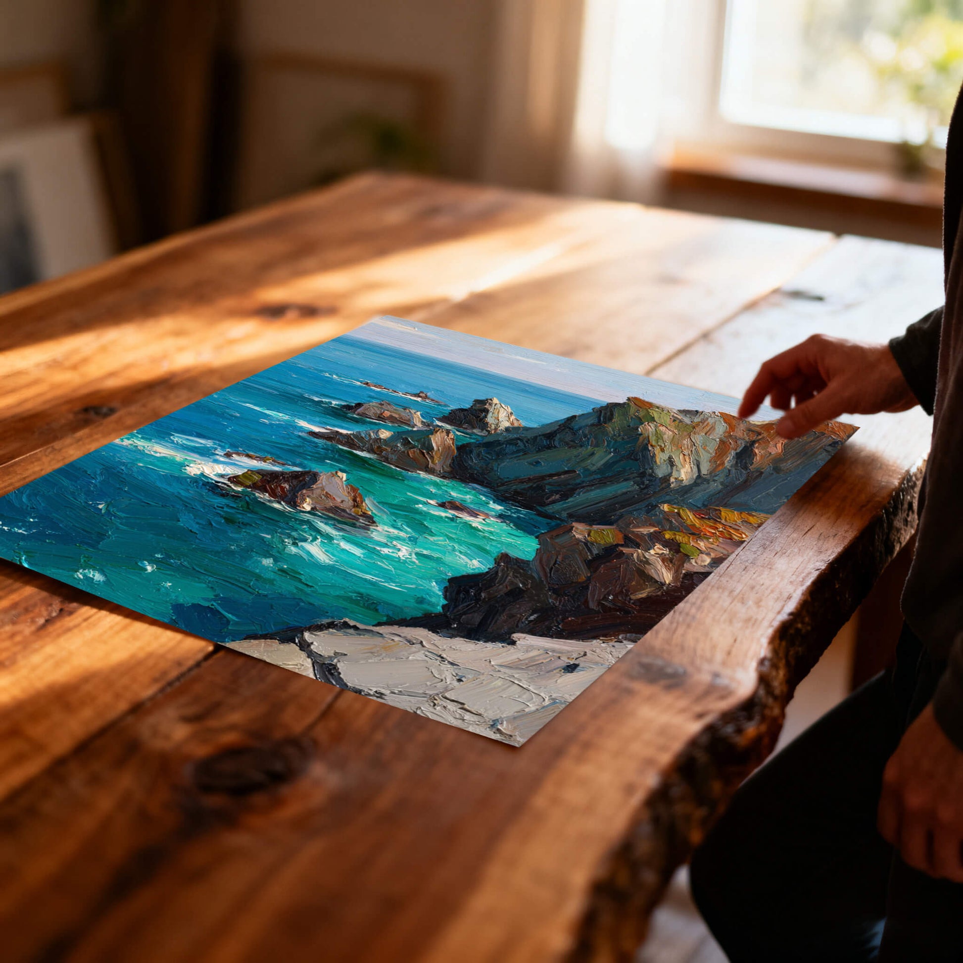 A close-up of the unframed seascape art print lying flat on a rustic wooden table, showing a person's hand reaching toward the textured print near a sunlit window.