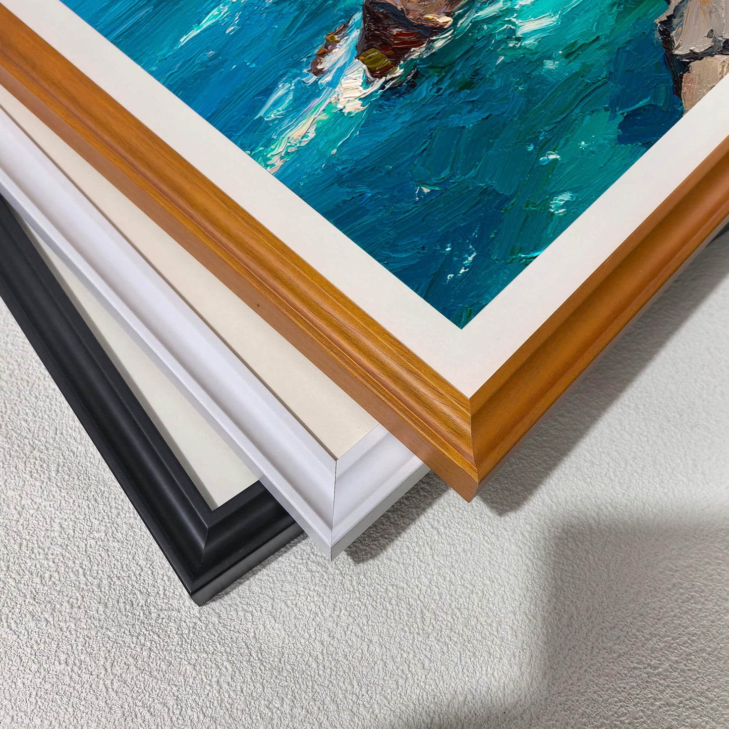A corner close-up showing the textured seascape artwork and three stacked frame options: warm wood, white, and black, demonstrating the available wood frame styles with a white mat.