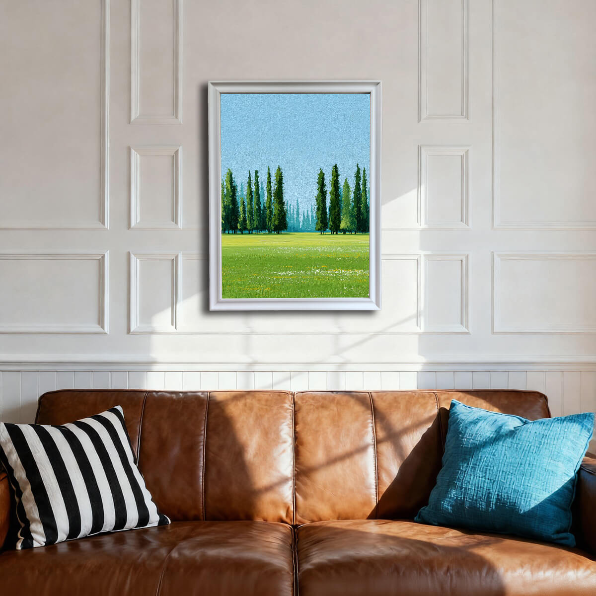 A framed art print of a bright green meadow with tall, slender Cypress trees against a light blue sky. The print is in a white frame, hanging above a brown leather sofa with a black/white striped pillow and a blue throw pillow in a room with white wainscoting.