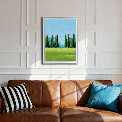 A framed art print of a bright green meadow with tall, slender Cypress trees against a light blue sky. The print is in a white frame, hanging above a brown leather sofa with a black/white striped pillow and a blue throw pillow in a room with white wainscoting.
