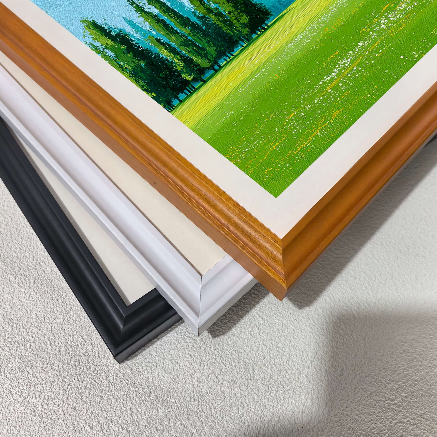 A stack of wood picture frame corners showing different color options: natural wood, cream/off-white, white, and black. A corner of the green meadow art print is visible on top.