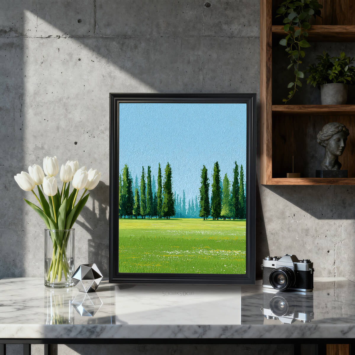 A framed art print of a bright green landscape with tall Cypress trees under a blue sky. The print is in a black frame, displayed on a marble-top table next to white tulips and a vintage camera in a modern, concrete-walled room.