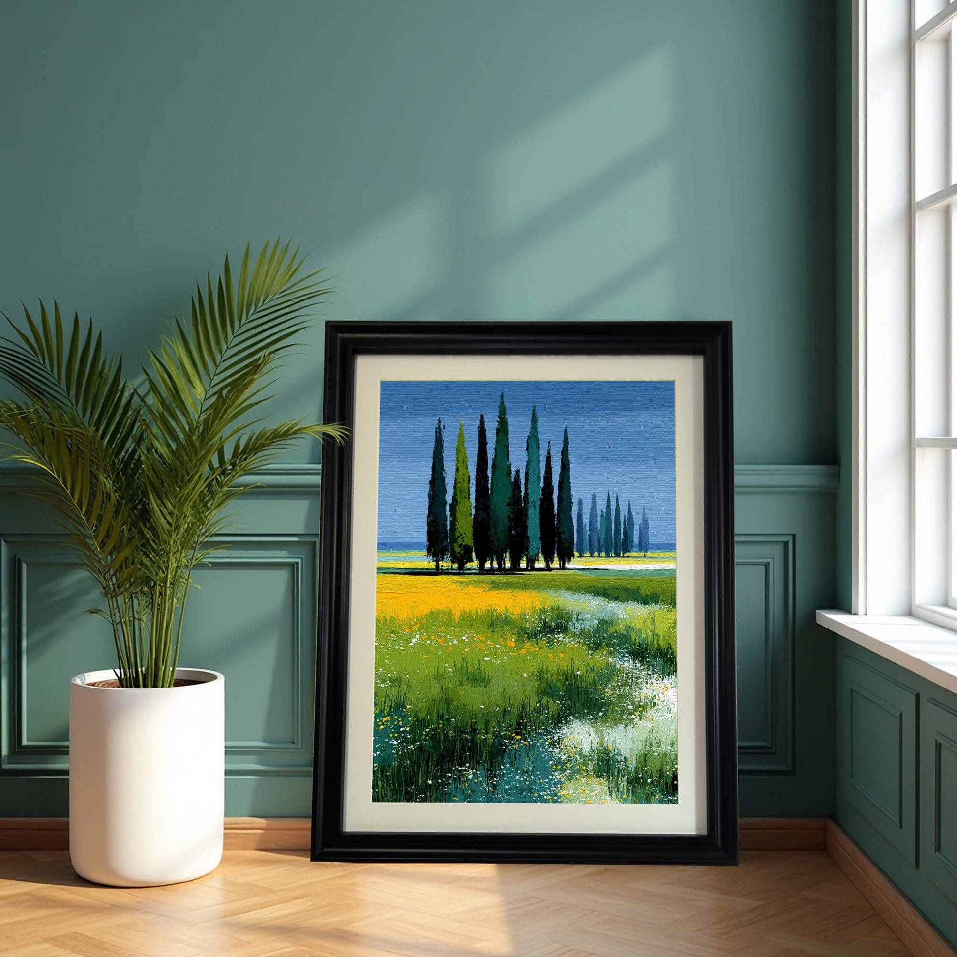 Art print of cypress trees in a field with a blue sky, framed in black, leaning on a wooden floor against a dark teal paneled wall next to a large potted palm.