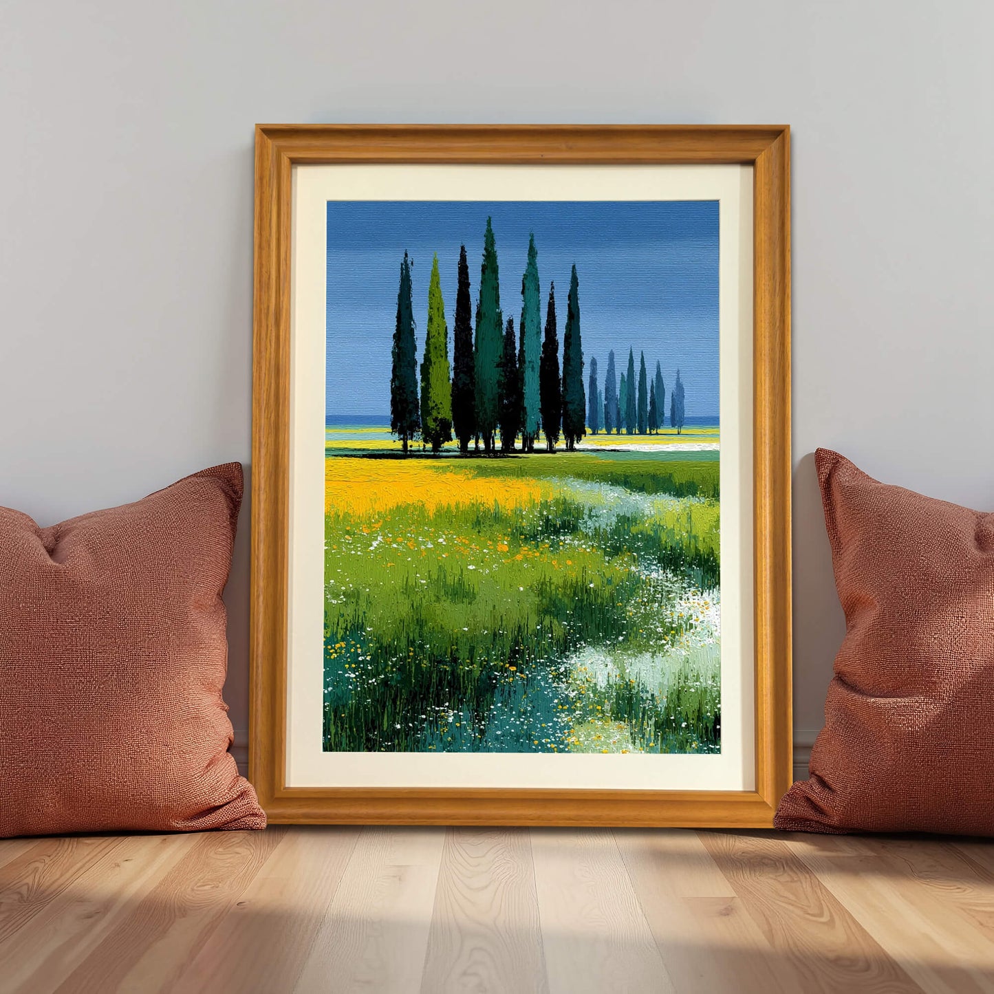 Framed art print of the cypress trees in a wildflower field in a light wood (natural) frame, leaning against a gray wall between two rust-colored pillows.