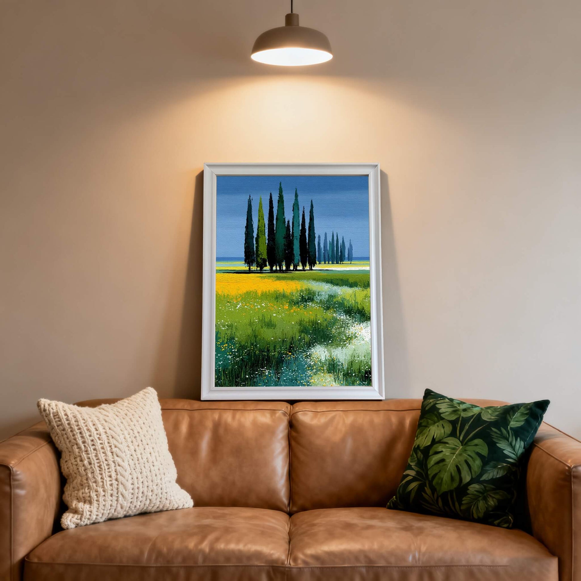 Art print of Tuscan cypress trees in a wildflower field, framed in white, leaning on a leather sofa with textured and tropical print pillows beneath a ceiling light.