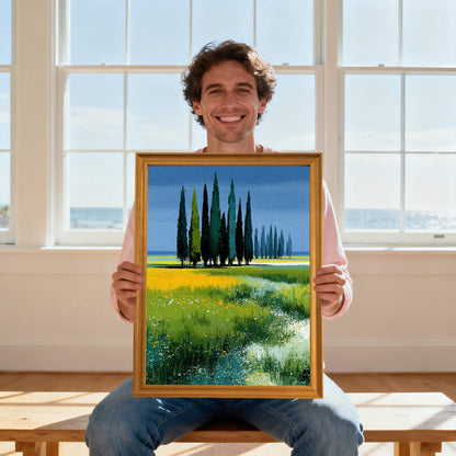 A smiling man holding up the art print of cypress trees in a yellow and green field, framed in light wood (natural), in front of a sunny window with an ocean view.