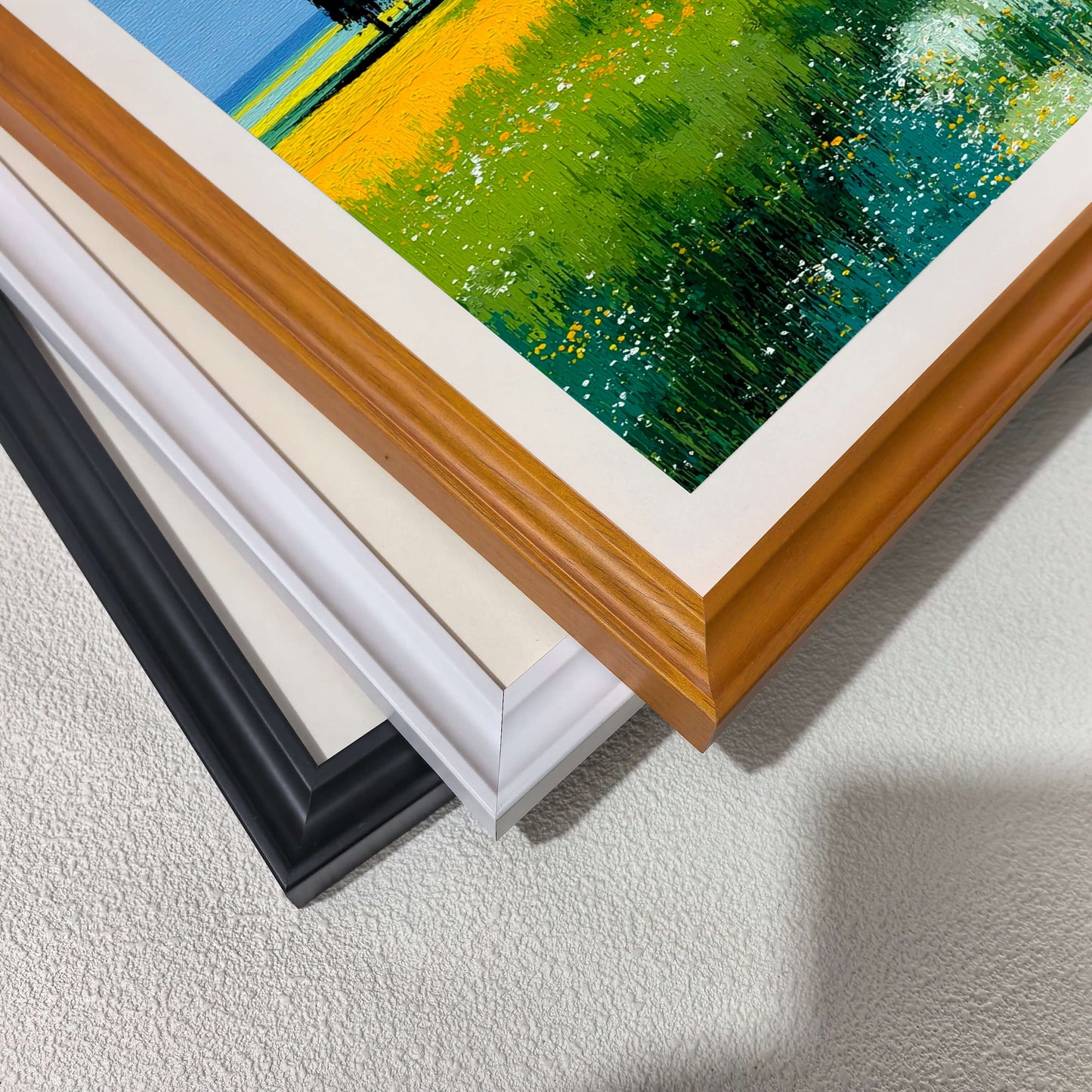 Stacked close-up view of the four different wood frame options: black, white, light wood (natural), and dark wood (brown/natural), with a corner of the vibrant green and yellow print visible.