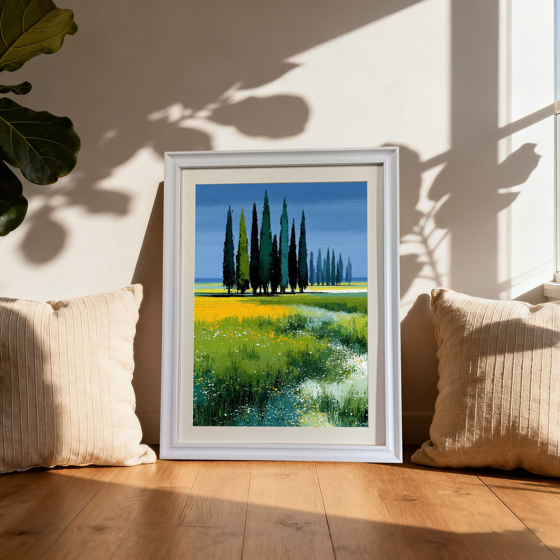 Framed print of cypress trees and a colorful meadow in a white frame, leaning on a wooden floor between two neutral pillows with natural sunlight and plant shadows.