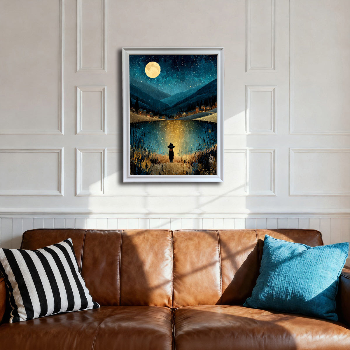 A white-framed art print centered above a brown leather sofa with striped and blue pillows. The white paneled wall provides a bright contrast to the dark, mystical nighttime landscape of the artwork.