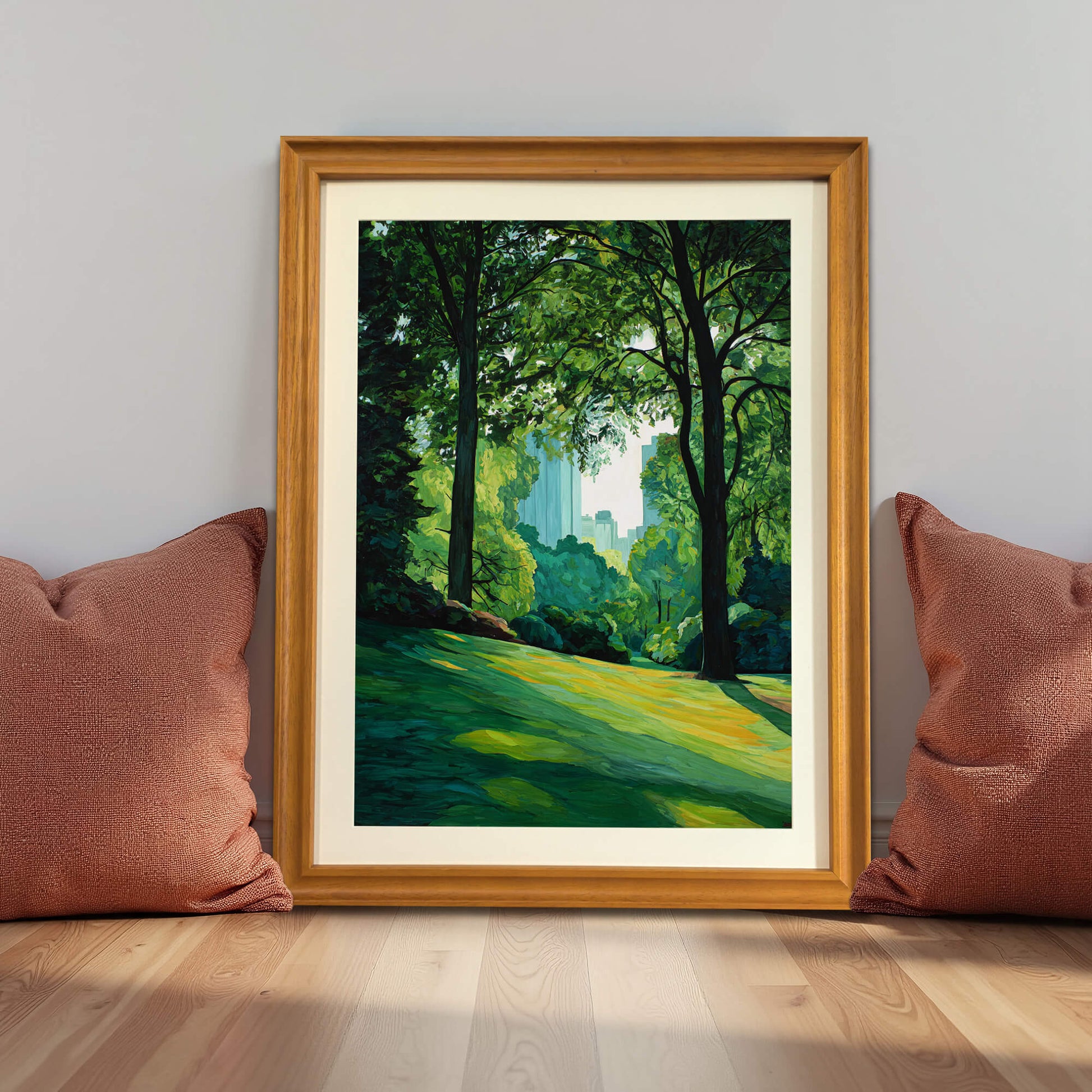 City park landscape art print in a natural light wood frame (oak/maple), leaning against a gray wall on the floor between two terracotta-colored pillows, demonstrating lifestyle decor.