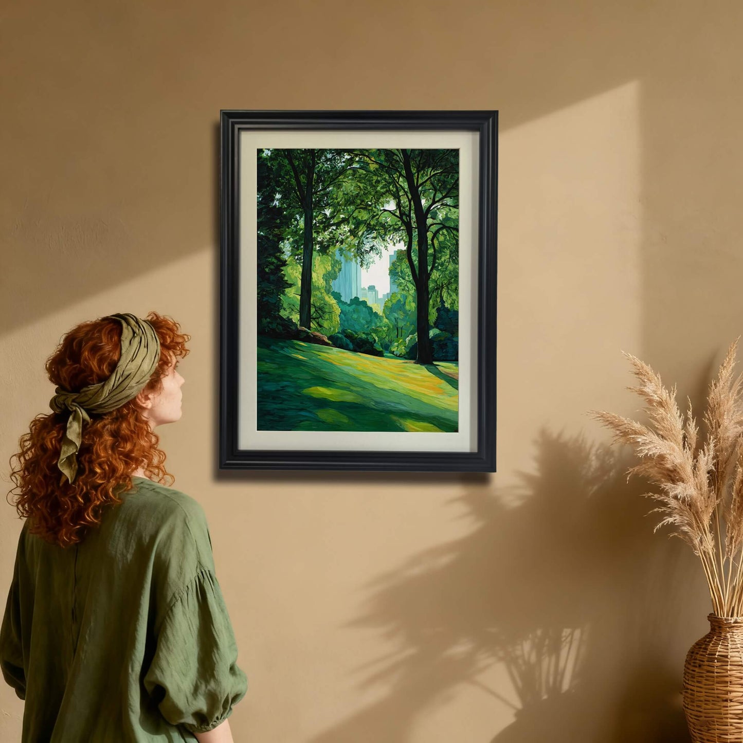 City park landscape art print in a black wood frame hanging on a tan wall. A woman in a green shirt is looking at the painting, showing the scale and the artwork's impact in a home setting.