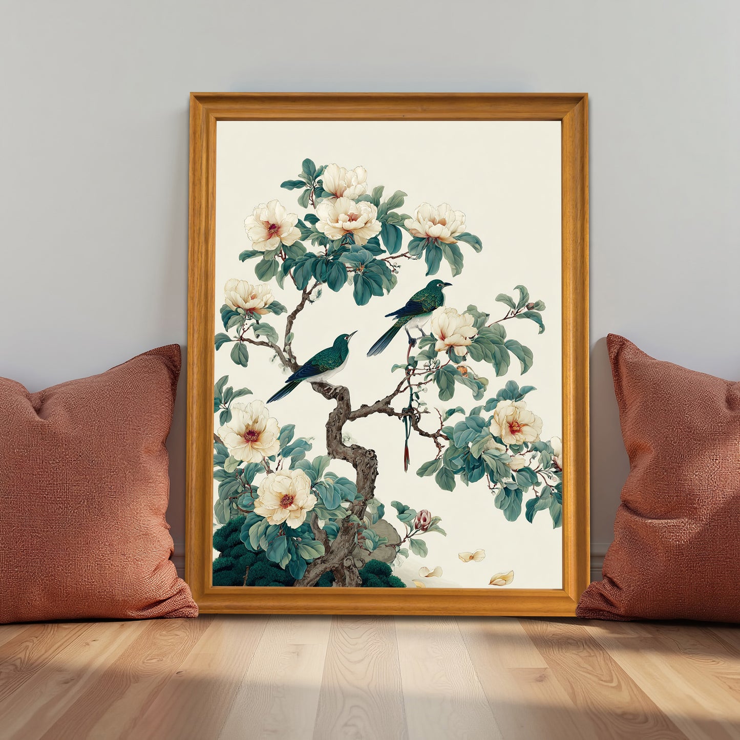A wooden framed botanical art print leaning against a white wall on a light wood floor, flanked by two reddish-brown textured pillows.