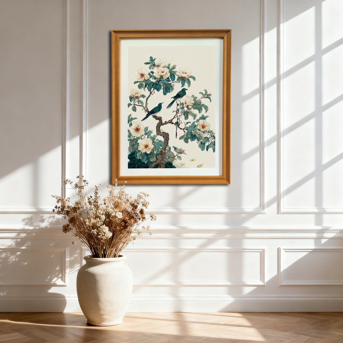 A light wood framed art print hanging on a white paneled wall above a large beige ceramic vase filled with dried flowers, with sunlight casting window shadows across the scene.
