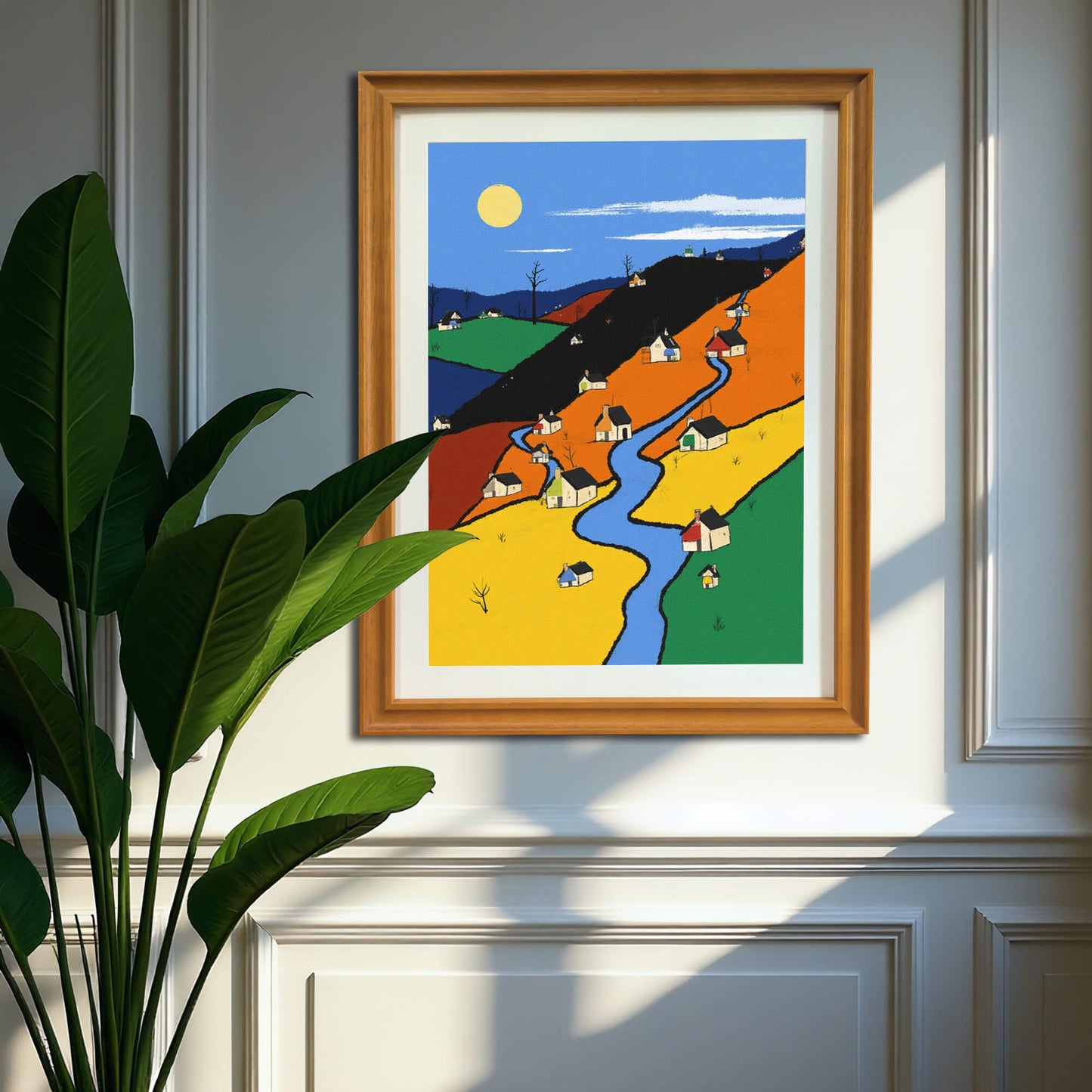 The color-block village art print, framed in warm wood, hanging on a light gray wainscoting wall, partially shadowed by a large, potted houseplant.