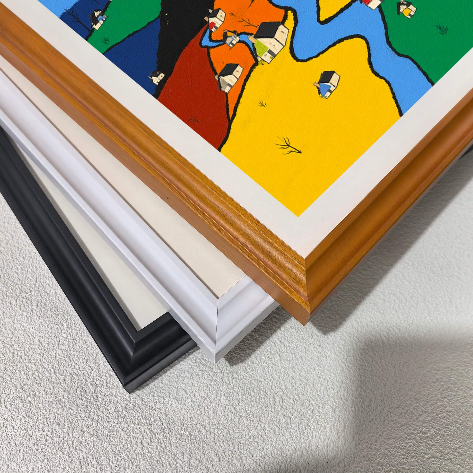A corner close-up showing the detail of the color-block village art print and three stacked frame options: warm wood, white, and black, demonstrating the available wood frame styles with a white mat.