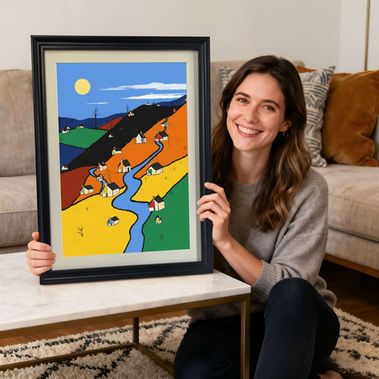 A smiling woman sitting on the floor, holding the color-block village art print framed in black wood, in front of a couch with pillows, showcasing the artwork.