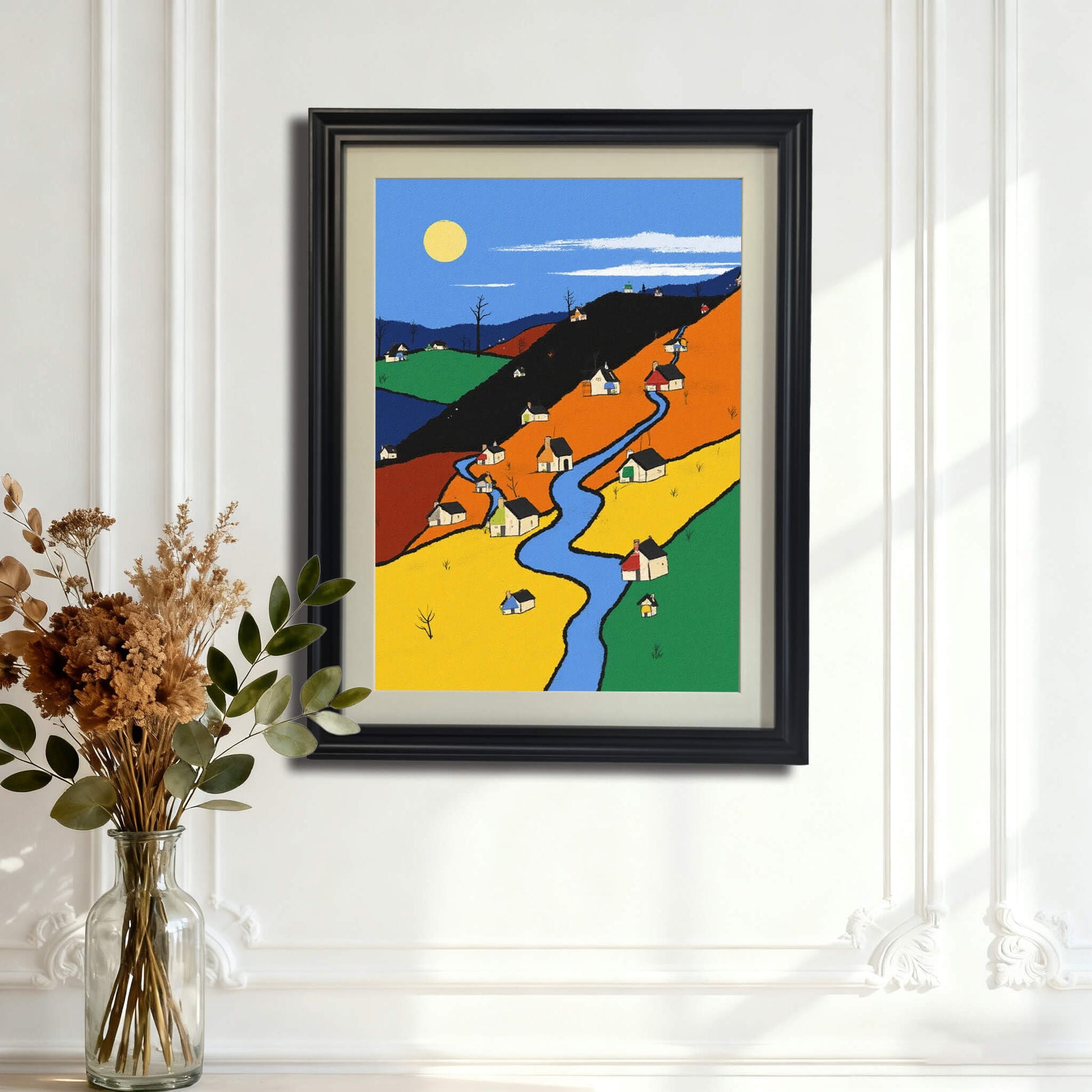 The color-block village art print, framed in black wood, hanging on a white wainscoting wall above a table with a clear vase of dried flowers and greenery.
