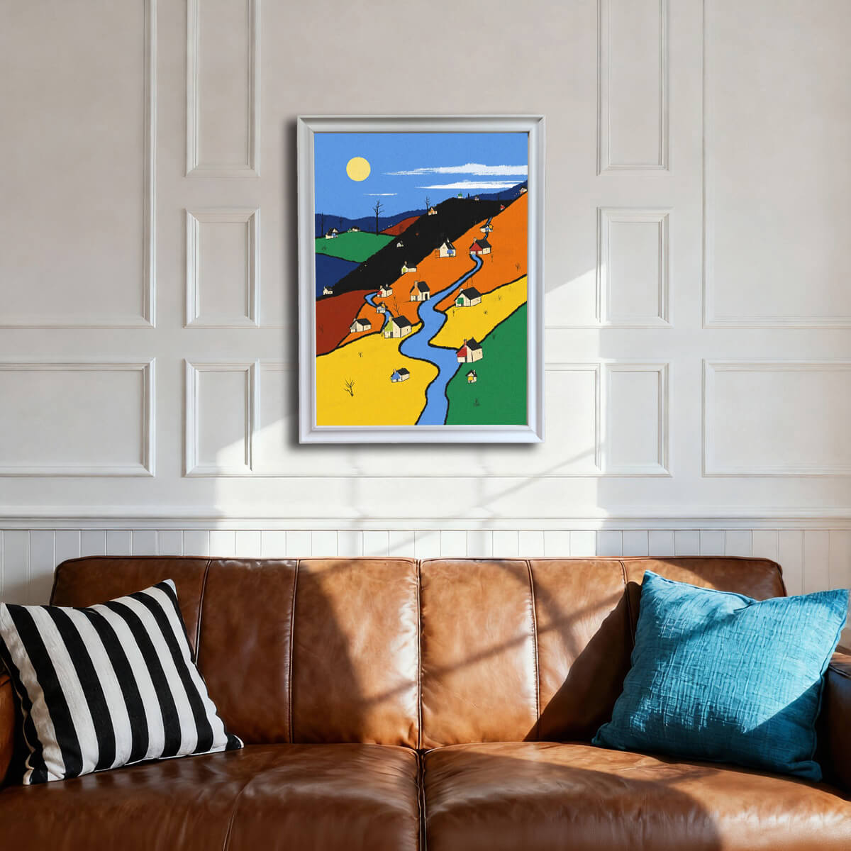 The color-block village art print, framed in white, hanging on a white paneled wall above a rich brown leather couch, flanked by black-and-white striped and teal throw pillows.