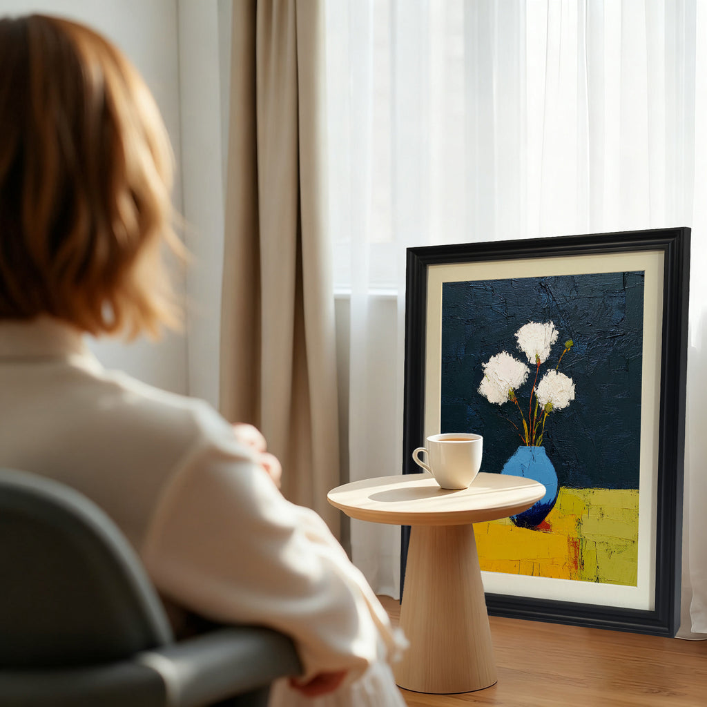 A black framed art print of white flowers in a blue vase, next to a small wooden table with a white teacup, in a sunlit interior with curtains.